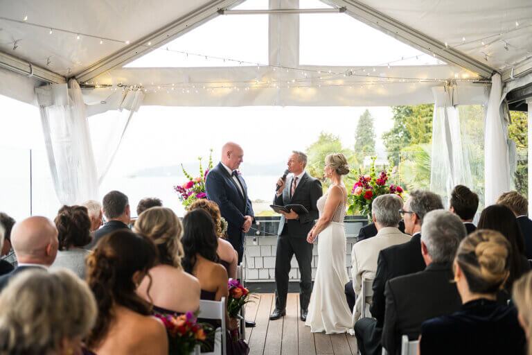 A wedding ceremony beneath a flower-draped tent with a waterfront view